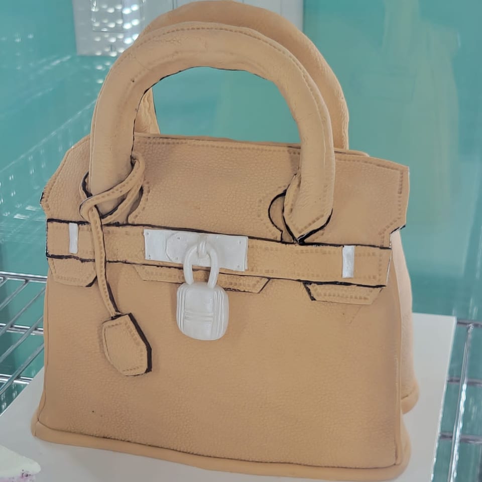 A beige handbag-shaped cake with white buckle and lock details sits on a white shelf.